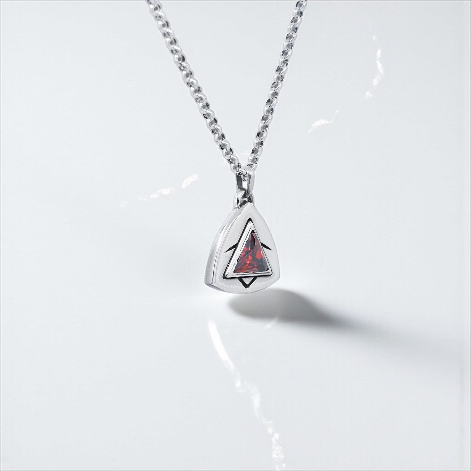 Minimalist silver pendant necklace with triangle-cut red birthstone, photographed in natural daylight to showcase the intricate hand-polished surface, ideal as a romantic anniversary or birthday gift for him or her.