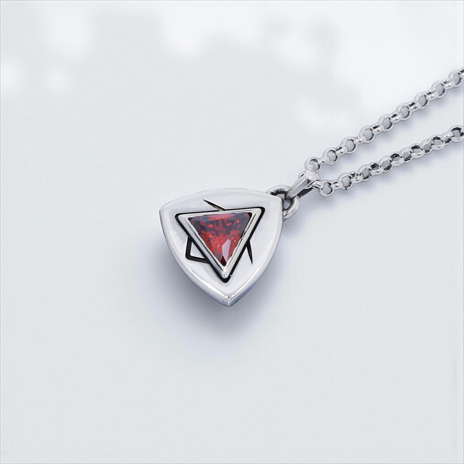 Close-up image of a handcrafted sterling silver pendant necklace featuring a vibrant triangle-cut ruby red gemstone at the center, set in a minimalist geometric bezel with sharp black accents, photographed on a clean white background to highlight the brilliance and artisan quality.