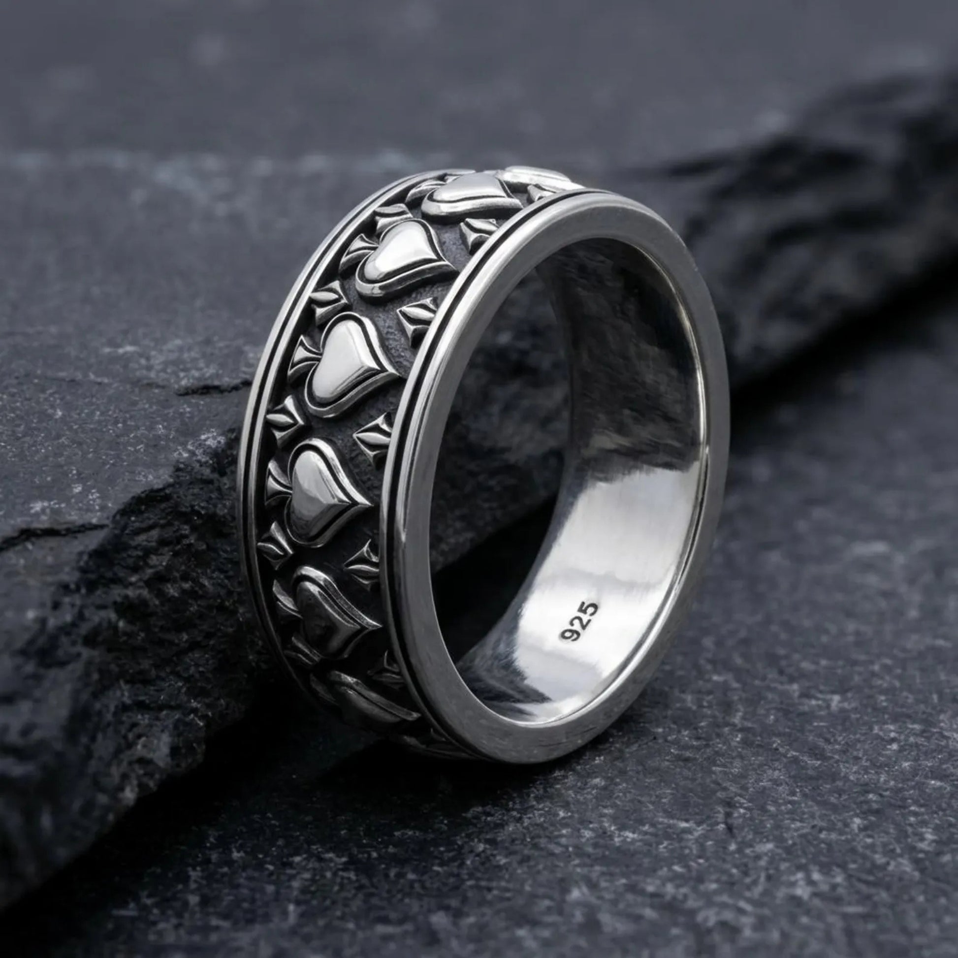 May include: A silver-colored ring featuring a repeating pattern of spade symbols around the outer band. The interior of the band is smooth and clearly stamped with the number 925, indicating the metal composition of the jewelry piece.