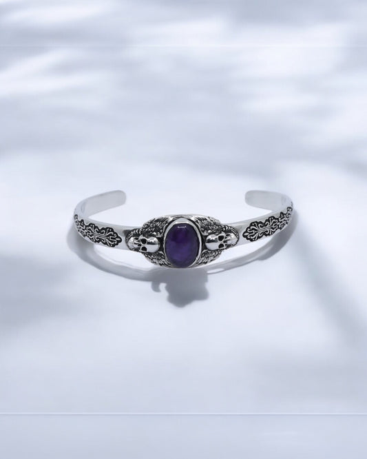 Close-up image of a handcrafted 925 sterling silver skull cuff bracelet featuring a deep purple amethyst gemstone in the center. The detailed gothic skull and wing engravings reflect premium craftsmanship and strength. Perfect for men who love handmade gothic jewelry and luxury silver accessories.