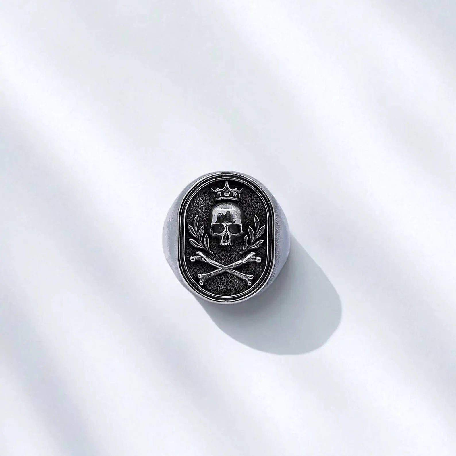 Top-down view showcasing the detailed skull and bone motif set within an oxidized textured oval frame, adding dramatic contrast and depth. The crown and laurel add a touch of dark royalty, making this ring a true symbol of power and individuality.