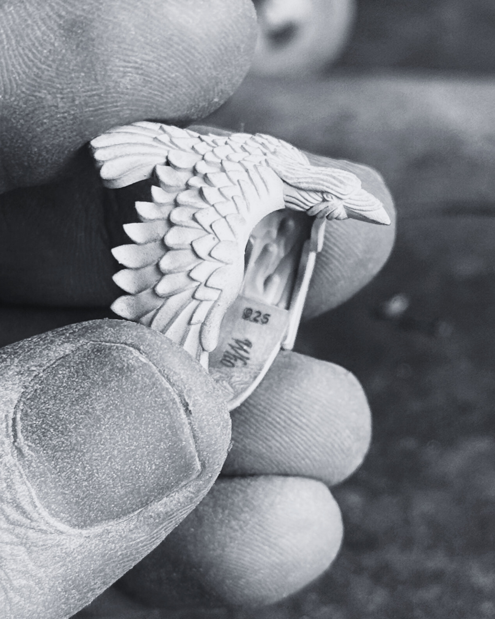 May include: A detailed, white metal bird sculpture, possibly a brooch or pendant, held in a hand. The bird has intricate feather detailing and a curved wing. The background shows tools and other metal pieces, suggesting a jewelry-making workshop.